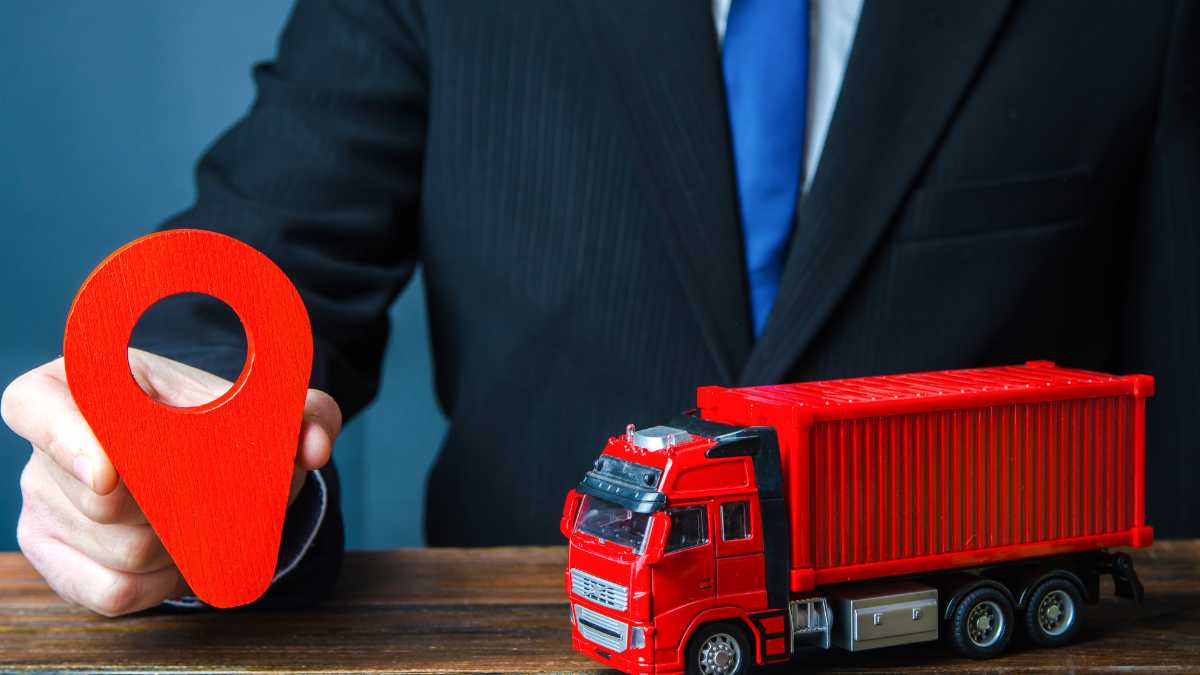 A hand holds a red pinpoint over a toy truck indicating logistics or location tracking in transport