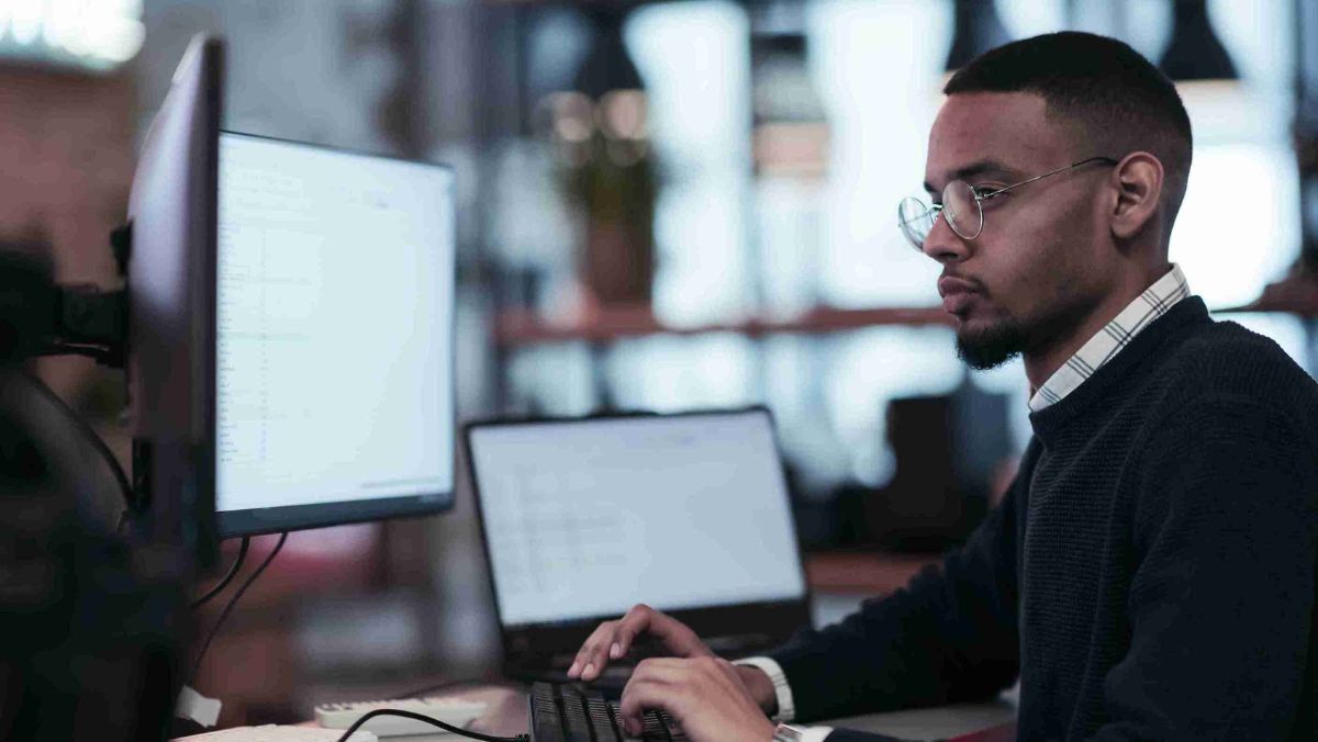 A man wearing eyeglasses is operating a computer with extended screens