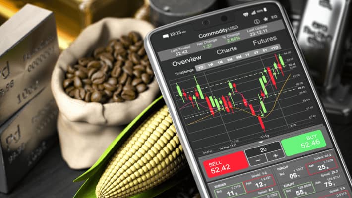 A smartphone displays commodity prices, surrounded by gold bars, coffee beans, corn, silver, and oil containers.