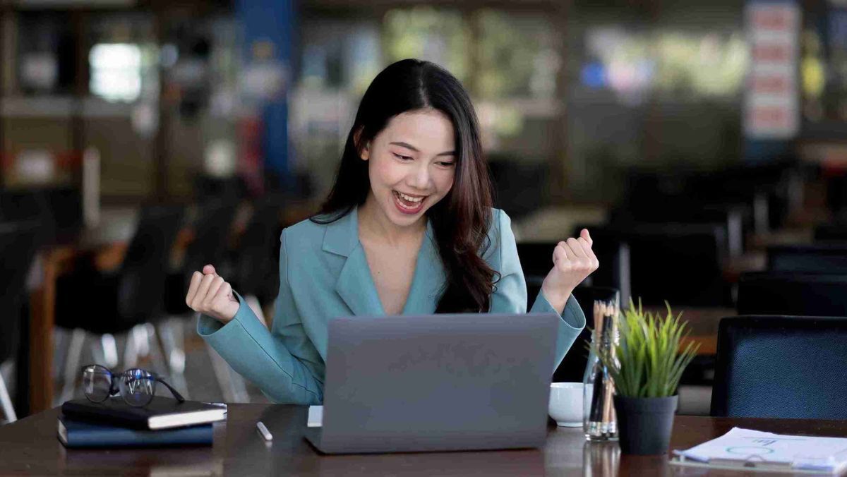 A woman in professional attire celebrating in front of her office laptop