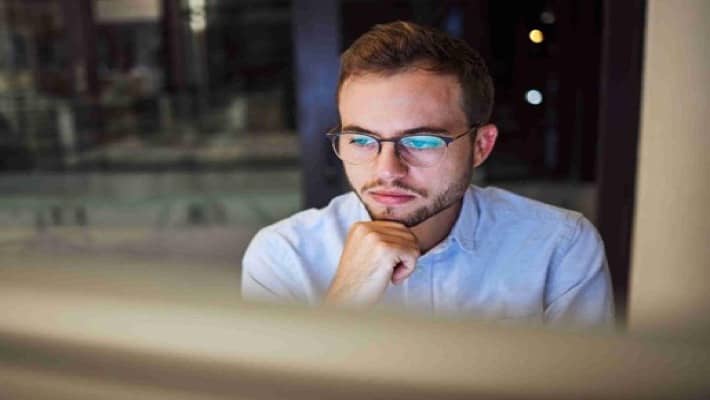 Man with glasses looking at a computer screen, deep in thought.