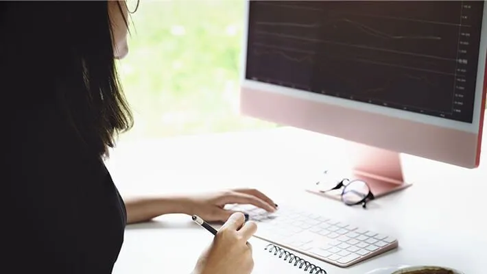 A woman analysing data on monitor while writing notes on a notepad