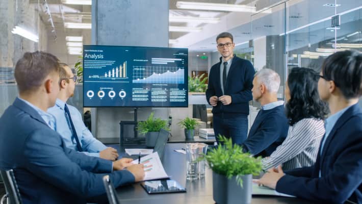 A group of professionals in a meeting room discussing, with one man standing and presenting to the team.