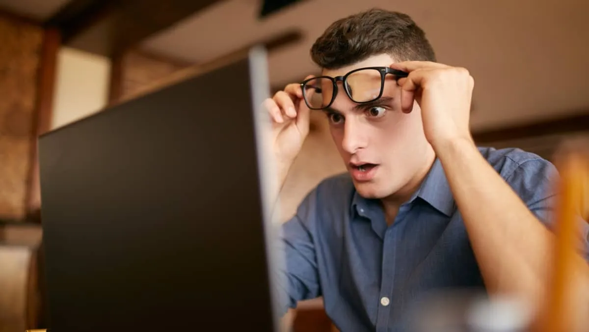 A man in disbelief lifting his eyeglasses and staring in shock at his laptop