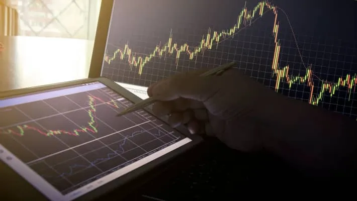 A hand with pen points to a tablet beside a monitor screen showcasing trading charts