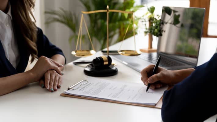 A person is signing a document across from another individual, with a gavel and scales of justice on the desk