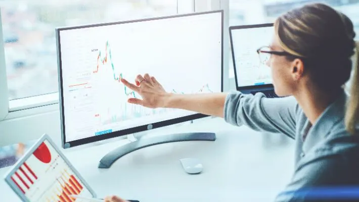 A woman analyses financial charts displayed across computer, tablet, and laptop screens