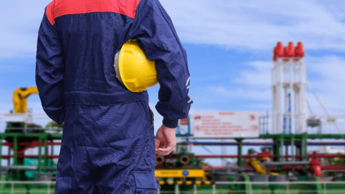 Worker in blue jumpsuit holding a yellow helmet, with industrial equipment in the background.