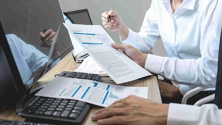 Two traders are sitting in front of a computer and examining financial documents